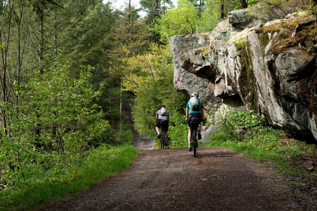 two people biking on a trail through the woods with rock on one side