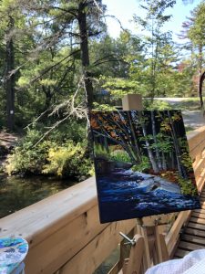 A artist's canvas sits in front of the forest landscape it represents; a bridge is partly visible
