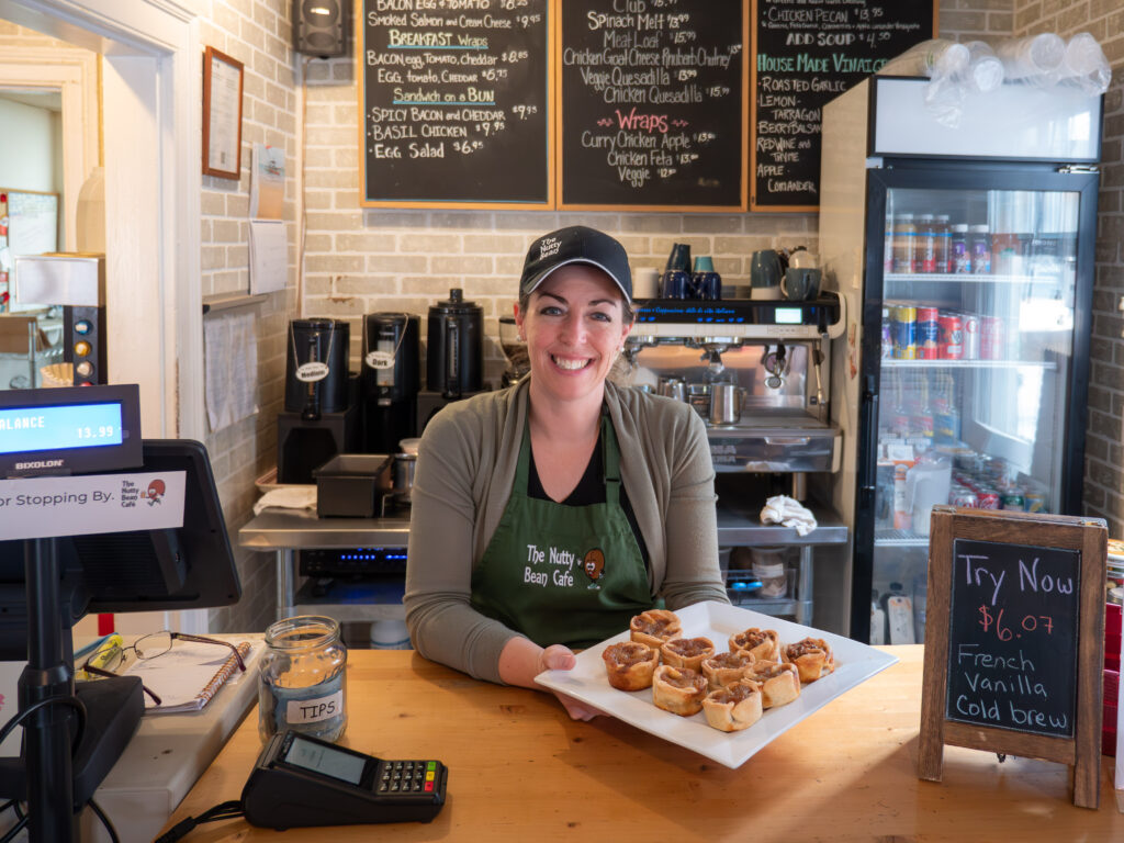 A smiling person behind a cafe counter holds a tray of butter tarts