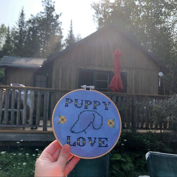Hand holding up a round embroidered sign that says puppy love in front of a cottage at Lake Edge Cottages in Lakesfield